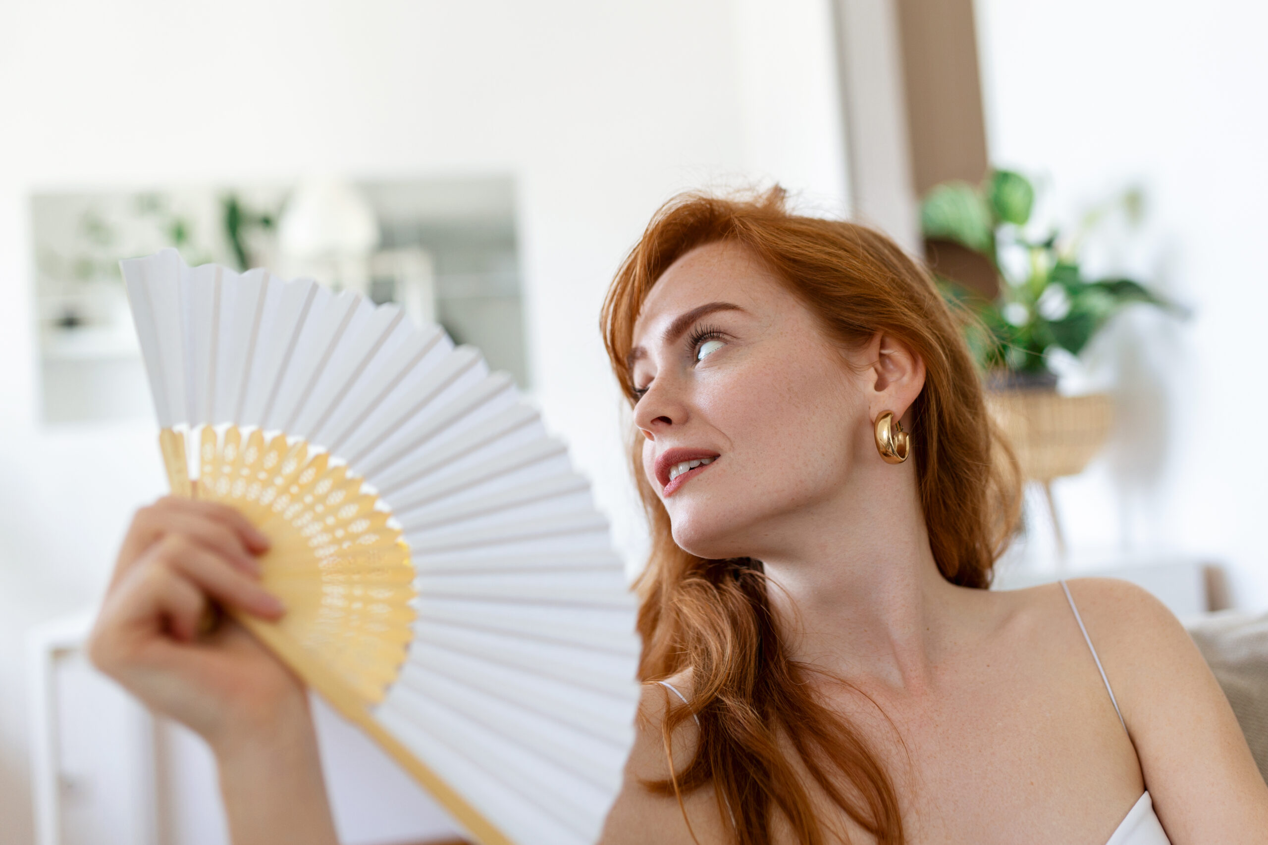 Head shot young woman using paper fan, feeling hot relieved indoors. Sweaty millennial cooling herself, breathing fresh air, spending time at home without conditioner.